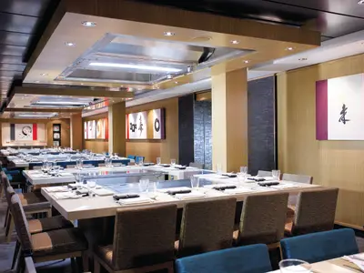 Modernes Teppanyaki Restaurant an Bord der Norwegian Epic. Lange Tische mit Kochplatten und gedecktem Geschirr. Elegantes Interieur mit Holzverkleidung und asiatischer Wandkunst.