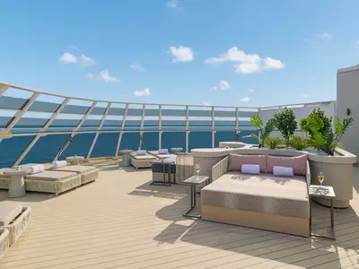 Ein modernes Sonnendeck an Bord der Norwegian Epic mit beigefarbenen Liegen und Sesseln, kleinen Tischen und grünen Pflanzen. Im Hintergrund ist das blaue Meer unter einem klaren Himmel zu sehen.