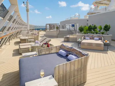 Das luxuriöse Sonnendeck der Norwegian Epic zeigt moderne Daybeds, Sonnenliegen und Beistelltische. Im Hintergrund sind ein Whirlpool, Pflanzen, das Meer und der blaue Himmel zu sehen. Die Szene ist entspannt.