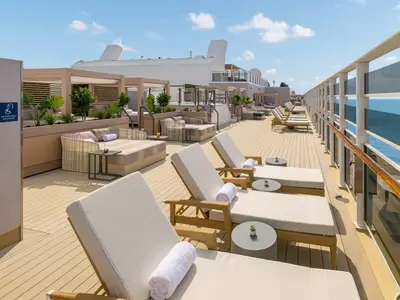 Weitläufiges Sonnendeck der Norwegian Epic mit beigen Liegen und Daybeds. Üppige Bepflanzung zwischen den Sitzbereichen. Rechts ist das blaue Meer sichtbar unter einem sonnigen Himmel.