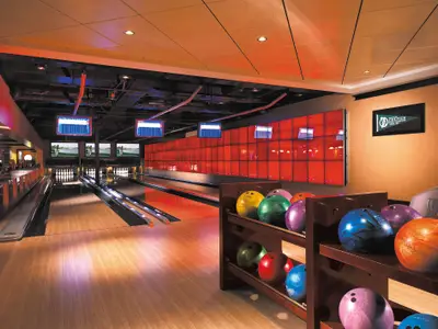 Eine helle Bowlingbahn an Bord der Norwegian Epic mit zwei Bahnen, Anzeigetafeln und einem Regal voller bunter Bowlingkugeln. Rote Wandverkleidung an der Seite.