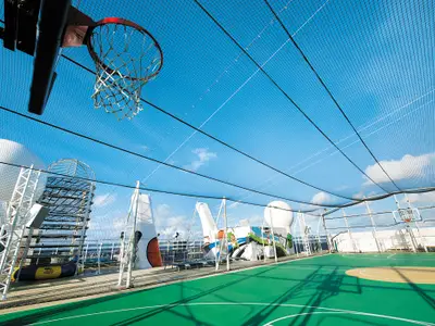 Weitwinkelaufnahme des grünen Basketballfelds an Bord der Norwegian Epic mit Sicherheitsnetz unter strahlend blauem Himmel. Zwei Körbe sind sichtbar. Im Hintergrund Strukturen des Schiffes.