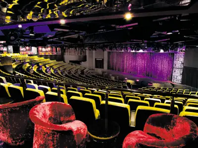 Uebersicht des geräumigen Theaters an Bord der Norwegian Epic mit zahlreichen gelben Sitzreihen und einer dramatisch beleuchteten violetten Buehne, ohne Publikum.