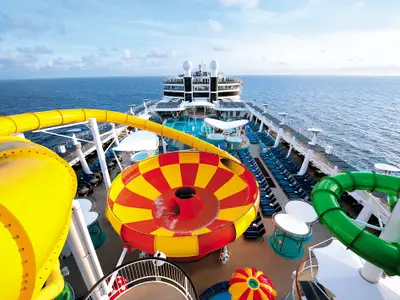 Vogelperspektive auf den Aqua Park der Norwegian Epic. Bunte Wasserrutschen, darunter rot-gelbe Trichterrutsche, auf Außendeck. Pools, Liegestühle, weites Meer unter blauem Himmel.