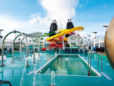 Panoramablick auf den Aqua Park der Norwegian Epic. Mehrere bunte Wasserrutschen, darunter eine gelbe Röhre, ein Pool und ein Spritzbereich mit Fontänen sind unter strahlend blauem Himmel zu sehen.