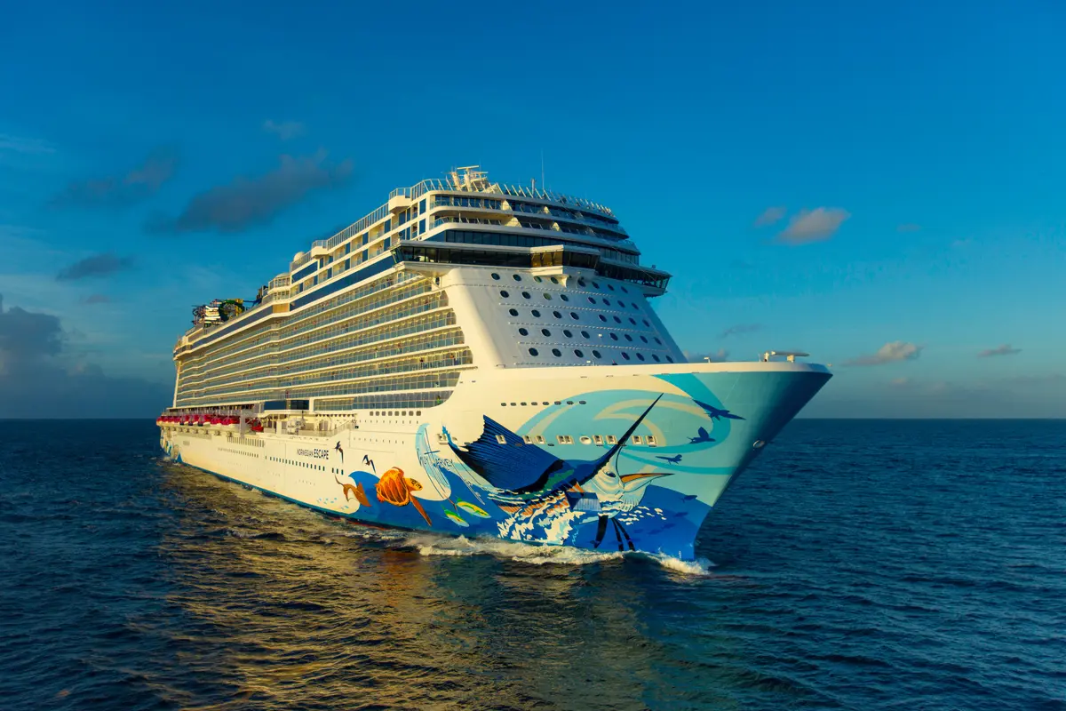 Die Norwegian Escape gleitet bei sonnigem Wetter durch das blaue Meer, von der Seite des Bugs aus gesehen. Die bunte Rumpfbemalung mit Meeresmotiven ist deutlich erkennbar.