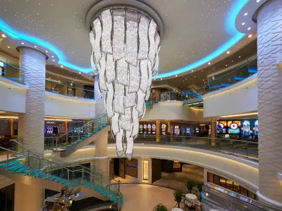 Das elegante, mehrstöckige Atrium an Bord der Norwegian Escape. Ein riesiger, funkelnder Kronleuchter, moderne Glas-Treppen, Shops und ein Casino-Bereich sind auf verschiedenen Ebenen zu sehen.