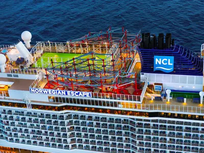 Luftaufnahme der Norwegian Escape bei Abenddämmerung. Auf dem Oberdeck sind ein Sportplatz, ein großer Kletterparcours und der Schornstein mit NCL-Logo zu sehen. Das Schiff fährt im Meer.