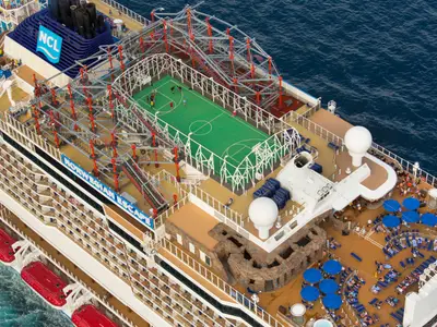 Vogelperspektive auf die belebten Oberdecks der Norwegian Escape mit Seilgarten, Sportplatz und Sonnendeck. Zahlreiche Passagiere geniessen die Tageszeit an Bord. Das Meer ist im Hintergrund sichtbar.