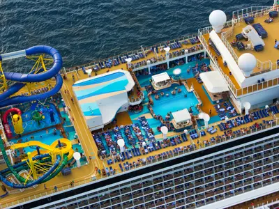 Draufsicht auf das belebte Pooldeck der Norwegian Escape mit mehreren Pools, bunten Wasserrutschen und zahlreichen Sonnenliegen, umgeben vom blauen Meer.