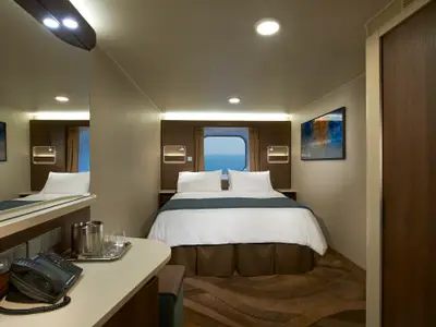 Eine helle, moderne Aussenkabine an Bord der Norwegian Escape mit einem Doppelbett und einem Bullaugenfenster, das den Blick auf das weite Meer freigibt. Ein Schreibtisch und ein Telefon sind ebenfalls zu sehen.