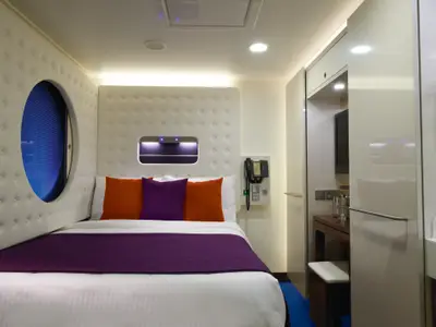 Helle Aussenkabine an Bord der Norwegian Escape. Ein Bett mit violetter Decke und bunten Kissen. Links ein Bullaugen-Fenster, rechts ein Schrank und Telefon.