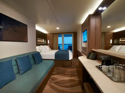 Helle und moderne Balkonkabine an Bord der Norwegian Escape. Zu sehen sind ein großes Bett, ein blaues Sofa, ein Fernseher, eine Kaffeestation und ein Balkon mit Meerblick.