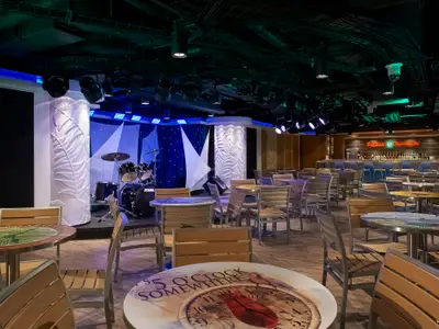 Ansicht der 5 O'Clock Somewhere Bar an Bord der Norwegian Escape. Eine beleuchtete Bühne mit Schlagzeug links, davor zahlreiche Tische und Stühle. Im Hintergrund die gut bestückte Bar.