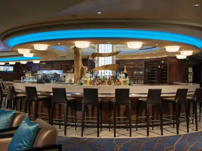 Das Atrium Bar & Cafe an Bord der Norwegian Escape zeigt eine helle, moderne Bar mit zahlreichen Flaschen und Barhockern. Die blaue Beleuchtung der Decke schafft eine einladende Atmosphäre.