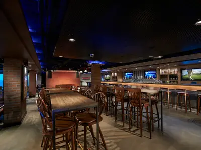 Geräumige Bar-Lounge an Bord der Norwegian Escape mit Holztischen, Barhockern und langer Theke. Blaue Akzentbeleuchtung und Backsteinwände schaffen ein modernes Ambiente. Bildschirme zeigen Sport und Specials.