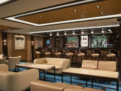 Eine modern eingerichtete Bar-Lounge an Bord der Norwegian Escape mit langer Bar, Barhockern, beigen Ledersofas und Tischen auf blauem Teppich. Die Bar ist reich bestückt mit Flaschen.