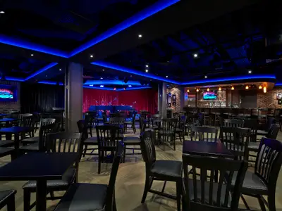 Innenansicht der Headliners Bar & Lounge an Bord der Norwegian Escape. Eine Bühne mit Klavier und rotem Vorhang, eine Bar mit Hockern sowie viele dunkle Tische und Stühle. Blaue Deckenbeleuchtung erhellt den Raum.