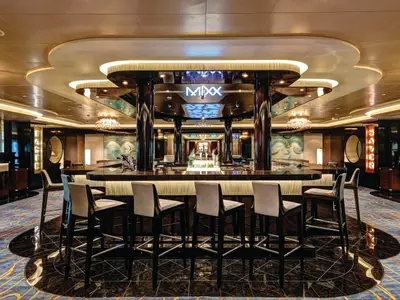 Die stilvolle MIXX Bar an Bord der Norwegian Escape, mit einem beleuchteten Barbereich, glänzenden Theken und bequemen Barhockern. Elegante Deckenbeleuchtung und gemusterter Teppichboden ergänzen das moderne Design. Die Bar ist leer.