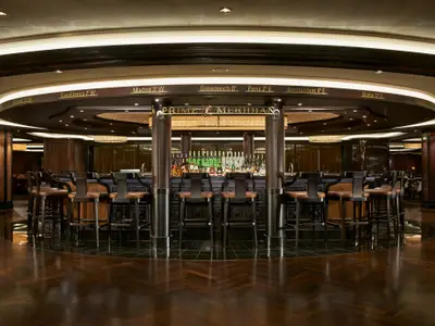 Die elegant eingerichtete Prime Meridian Bar an Bord der Norwegian Escape. Eine runde Bar mit dunklen Barhockern, umgeben von dunklem Holz und Lichtern. Zahlreiche Flaschen stehen hinter der Bar, und oben sind Städtenamen mit Längengraden zu sehen, die auf den Prime Meridian Bezug nehmen.