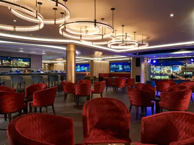 Skyline Bar an Bord der Norwegian Escape. Rote Sessel, Barhocker, Bildschirme mit nächtlicher Stadtansicht. Elegante runde Kronleuchter illuminieren den modernen Bar-Bereich.