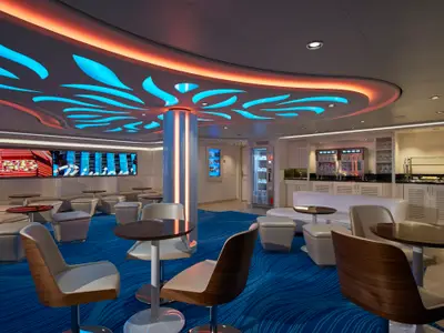 Die Bar-Lounge der Norwegian Escape zeigt moderne Einrichtung mit blauen und orangen Deckenleuchten. Sichtbar sind Sitzbereiche, ein Casino und eine Bar. Der Teppich ist blau.