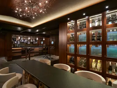 Elegante Bar-Lounge an Bord der Norwegian Escape, dominiert von dunklem Holz und einem verzweigten Kronleuchter. Beleuchtete Vitrinen präsentieren Weinflaschen. Im Vordergrund eine lange Theke mit hellen Hockern, dahinter ein Barbereich mit runden Fenstern. Gedämpfte Beleuchtung schafft eine gemütliche Atmosphäre.