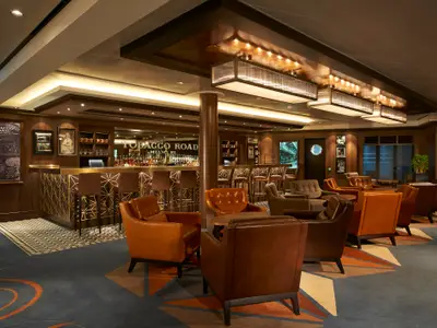 Elegante Tobacco Road Bar an Bord der Norwegian Escape. Holzbar mit Goldmuster, Flaschen und bequeme Ledersessel in einer gemütlichen Lounge. Stimmungsvolle Deckenleuchten.