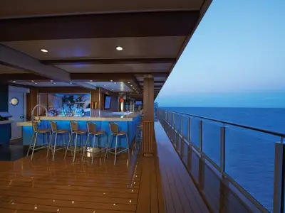 Die beleuchtete 5 o'Clock Somewhere Bar an Bord der Norwegian Escape bei Dämmerung mit Barhockern, blauer Beleuchtung und Meerblick auf der Steuerbordseite.