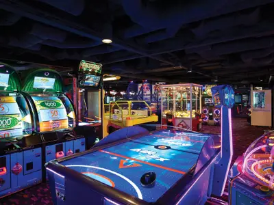 Bunte und lebhafte Spielhalle an Bord der Norwegian Escape. Ein Airhockey-Tisch und zahlreiche leuchtende Arcade-Automaten füllen den dunklen Raum mit Licht und Farbe. Eine dynamische Unterhaltungszone ohne Personen.