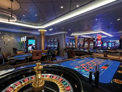 Das Casino der Norwegian Escape zeigt einen blauen Roulette-Tisch im Vordergrund, flankiert von Spielautomaten und weiteren Spieltischen im Hintergrund. Die Decke ist gemustert und mit eleganten Lichtleisten versehen.