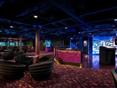 Club Entourage auf der Norwegian Escape: Gaming-Stationen, violette Bar, schwarze Lounge-Sessel, Graffiti-Wandkunst. Moderne Neonlichter schaffen eine lebhafte Atmosphäre.