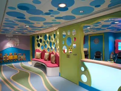 Ein farbenfroher Kinderbereich an Bord der Norwegian Escape. Die Wände sind hellblau und lila mit Tierillustrationen, die Decke ist weiß mit blauen Wolkenmustern. Ein gelb-grüner Spielbereich mit runden Öffnungen und einer rosafarbenen Sitzbank ist sichtbar.