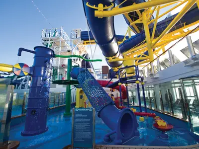 Der Kids AquaPark an Bord der Norwegian Escape zeigt bunte Wasserrutschen, eine Wasserspielstruktur mit Eimer und Spritzelementen unter blauem Himmel. Das Deck ist nass und glänzt in der Sonne.