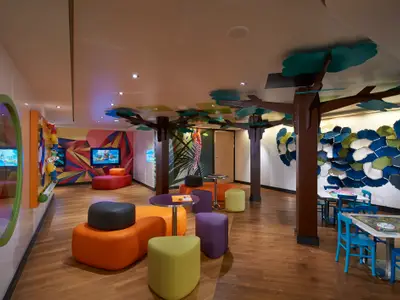 Bunter Kinderbereich an Bord der Norwegian Escape mit Spielgeräten, Bildschirmen, farbigen Sitzmöbeln und Baumdekorationen.