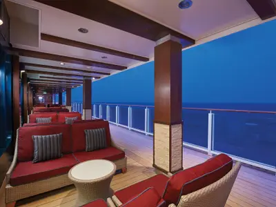 Panoramablick auf die überdachte Waterfront Lounge an Bord der Norwegian Escape in der blauen Stunde. Bequeme rote Sofas stehen auf einem Holzdeck mit Blick auf das Meer und den Horizont.