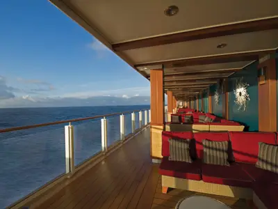 Blick über eine lange, überdachte Promenade auf der Norwegian Escape mit roten Loungesofas, türkisblauer Wandkunst und einem Glasgeländer zum offenen Meer.