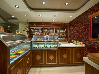 Detailansicht des Bake Shops an Bord der Norwegian Escape. Man sieht Theken mit Vitrinen voller bunter Macarons, Cupcakes und Glaeser mit Suessigkeiten. Die Waende sind mit braunen Kacheln und Holzregalen verziert.