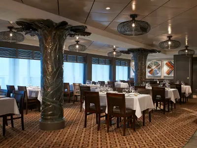 Elegantes Restaurant Bayamo auf der Norwegian Escape. Gedeckte Tische, dunkle Stühle, baumartige Säulen und Hängelampen sind sichtbar.