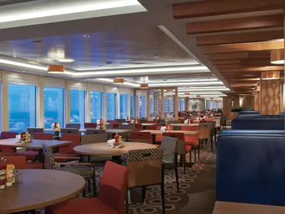 Geräumiges Restaurant an Bord der Norwegian Escape. Zahlreiche Holztische, rote Stühle, blaue Sitzbänke. Große Fensterfront mit Meer- und Stadtblick. Tische sind vorbereitet.
