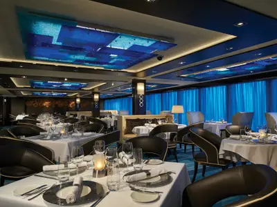 Elegantes Restaurant an Bord der Norwegian Escape. Weiße Tische, dunkle Sessel, blaue Deckenbeleuchtung und Vorhänge erzeugen maritime Atmosphäre. Keine Personen.