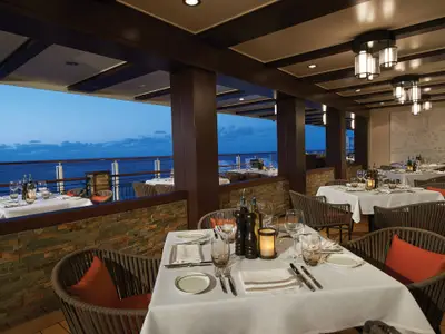 Blick in ein stilvolles Spezialitätenrestaurant an Bord der Norwegian Escape bei Abenddämmerung mit Tischen, Stühlen und Panoramablick auf das Meer.