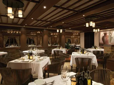 Blick in das elegant eingerichtete Spezialitätenrestaurant La Cucina an Bord der Norwegian Escape. Zahlreiche Tische sind weiß eingedeckt, umgeben von geflochtenen Stühlen. Im Hintergrund sind kunstvolle Fensterverzierungen und eine beleuchtete Wandmalerei zu sehen. Die Decke ist mit dunklen Holzbalken verziert und moderne Leuchten spenden warmes Licht.