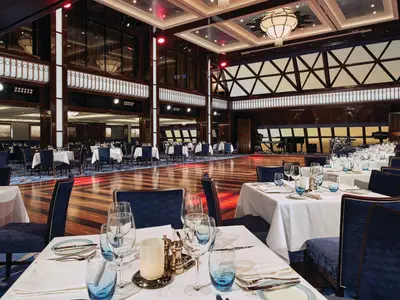 Blick in den grossen und eleganten Speisesaal Manhattan Room an Bord der Norwegian Escape mit zahlreichen weiss gedeckten Tischen, blauen Stühlen, einer grossen Tanzfläche aus Holz und einer Bühne mit Musikinstrumenten im Hintergrund. Deckenbeleuchtung und rotes Akzentlicht erzeugen eine festliche Abendstimmung.