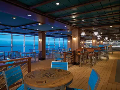 Ein helles Restaurant an Bord der Norwegian Escape mit Tischen, Stühlen und einer Bar. Große Fenster bieten einen weiten Blick auf das blaue Meer.