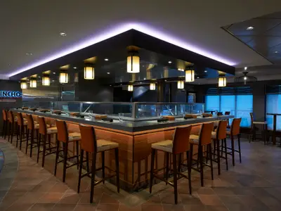 Blick in das stilvolle Tapas-Restaurant Pincho an Bord der Norwegian Escape mit langer Bar, hohen Stühlen und indirekter Beleuchtung. Fenster mit blauem Licht im Hintergrund.