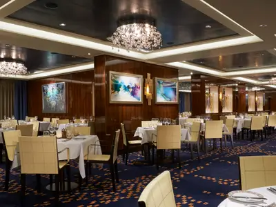 Elegantes Restaurant Savor an Bord der Norwegian Escape. Gedeckte Tische, beigefarbene Stühle, dunkle Holzpaneele, Kunstwerke, Kronleuchter.