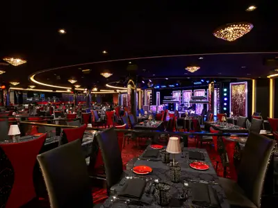 Spezialitätenrestaurant 'Esc Supper Club' an Bord der Norwegian Escape. Elegante Tische, rote Stühle und eine Showbühne. Luxuriöses Ambiente mit stimmungsvoller Beleuchtung.