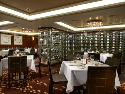 Elegantes Restaurant an Bord der Norwegian Escape. Gedeckte Tische, dunkle Stühle. Große Weinwand und gläserner Weinkeller im Hintergrund. Warme Beleuchtung schafft luxuriöses Ambiente.