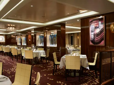Elegantes Restaurant an Bord der Norwegian Escape. Gedeckte Tische, cremefarbene Stühle, dunkle Holzvertäfelung mit Kunstwerken und blauen Skulpturen in Nischen. Roter Teppichboden.